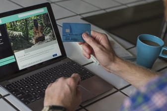 Ein Mann sitzt mit seiner Kreditkarte in der Hand vor einem Notebook: Zahlen Sie bei der Buchung einer Ferienunterkunft nicht sofort den Gesamtbetrag.