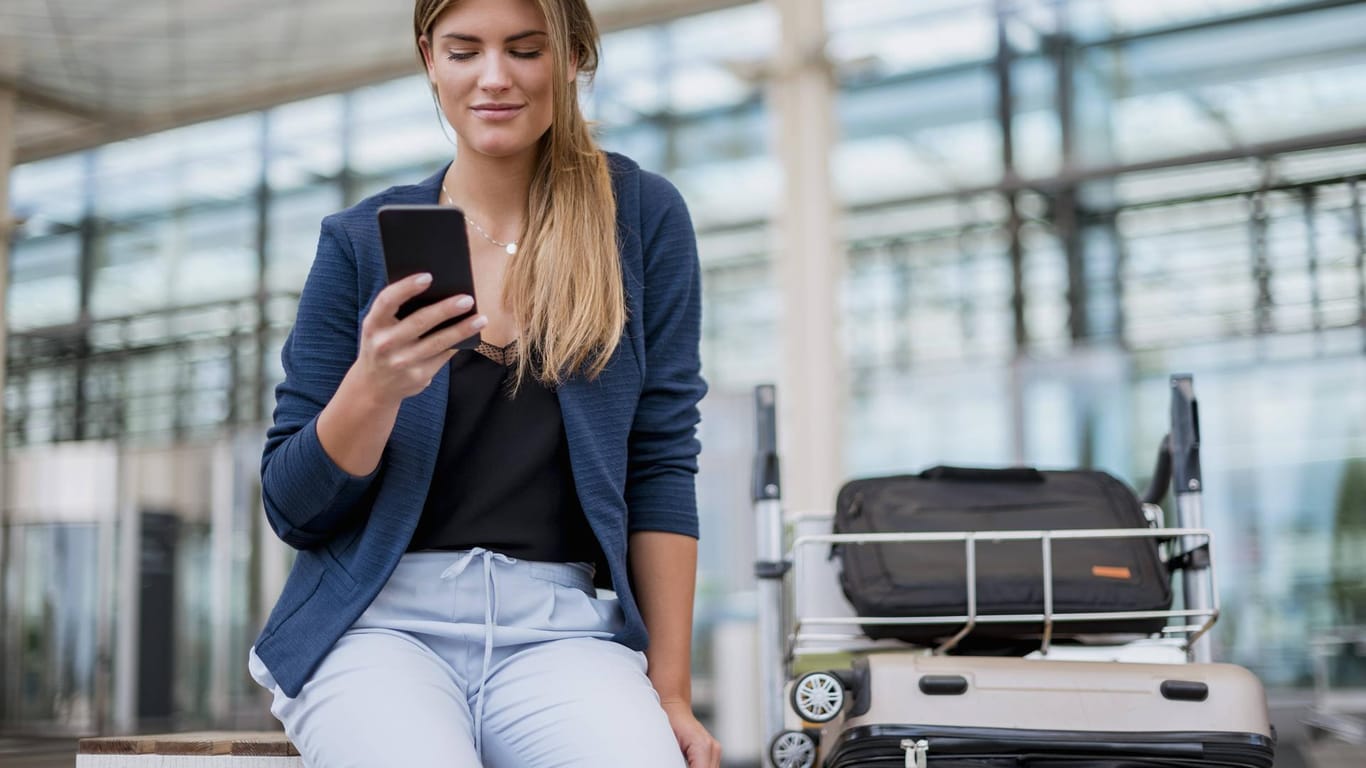 Eine junge Frau sitzt mit ihrem Gepäck am Flughafen und nutzt ihr Smartphone: Vor einer Reise sollte man eine digitale Kopie seines Ausweises anfertigen und abspeichern.