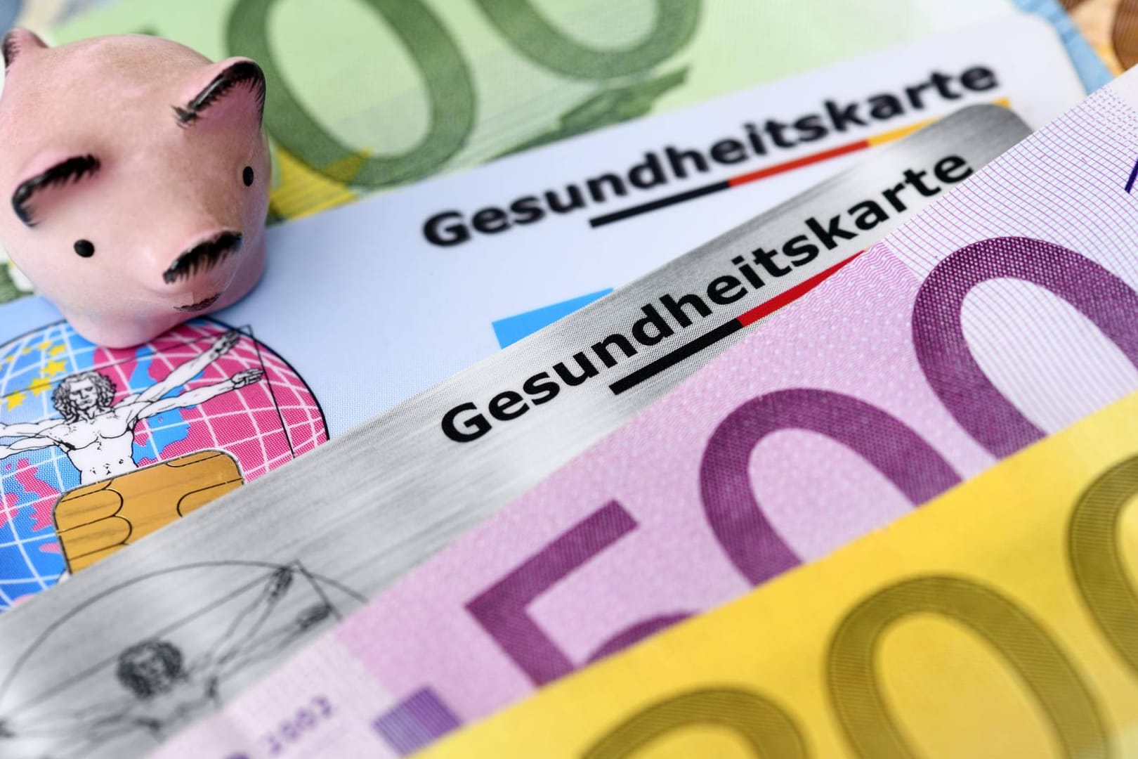 Gesundheitskarten mit Geldscheinen und Sparschwein: Bei den Krankenkassenbeiträgen lohnt sich auch der Vergleich mit regionalen Anbietern. (Symbolbild)