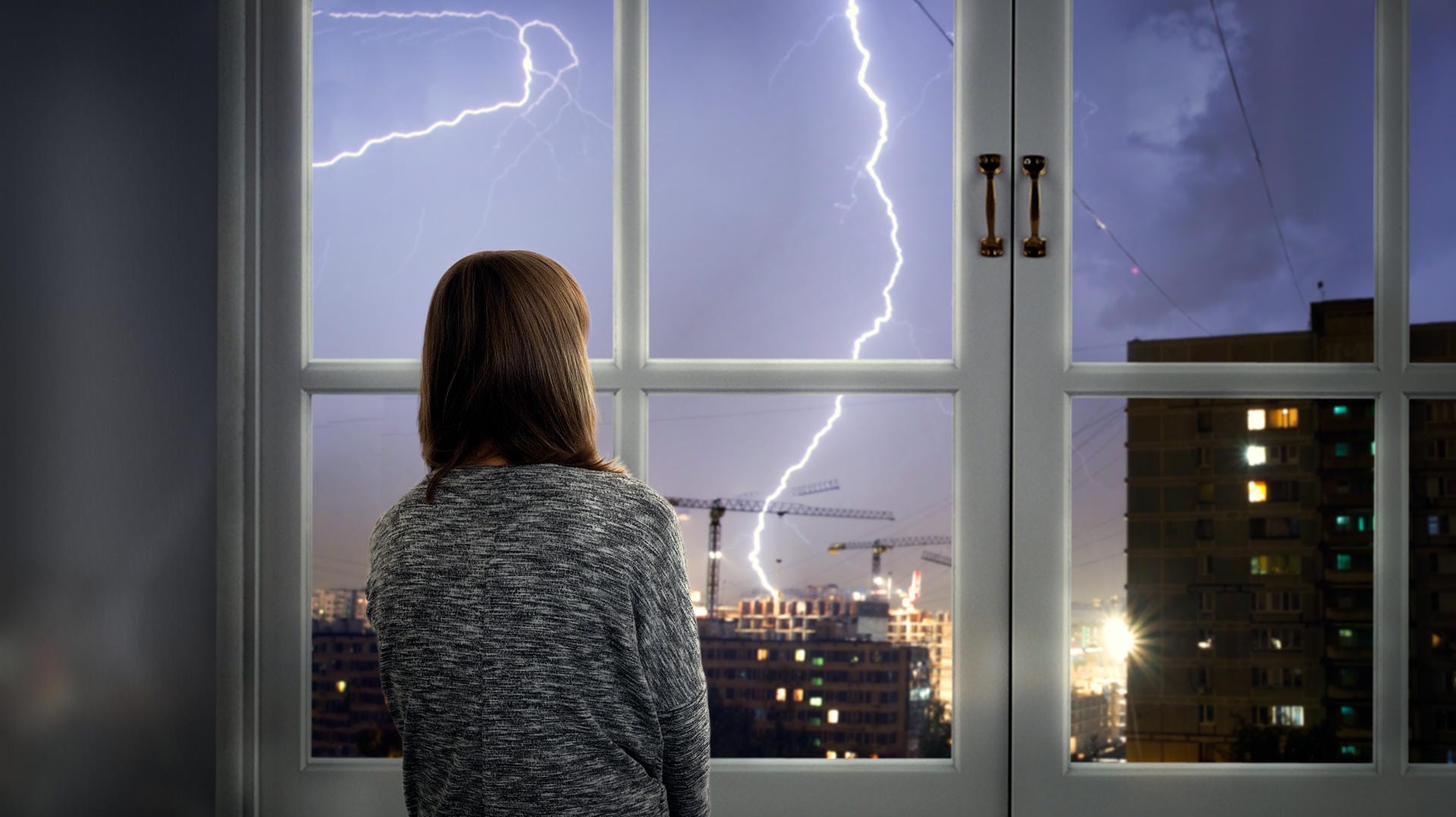 Eine junge Frau sieht sich ein Gewitter vom Fenster aus an: Haushaltselektronik sollte während eines Unwetters ausgesteckt werden.