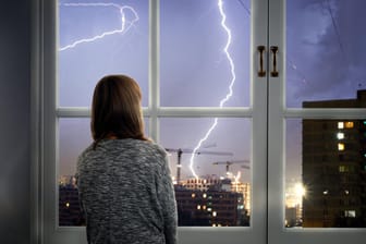 Eine junge Frau sieht sich ein Gewitter vom Fenster aus an: Haushaltselektronik sollte während eines Unwetters ausgesteckt werden.