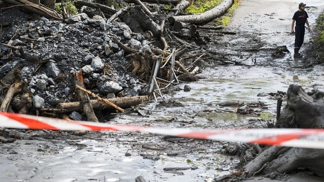 Ein Mann steht auf einer abgesperrten Straße, die von Schlamm, Bäumen und Geröll bedeckt ist. Ein Mann steht auf einer abgesperrten Straße, die von Schlamm, Bäumen und Geröll bedeckt ist.