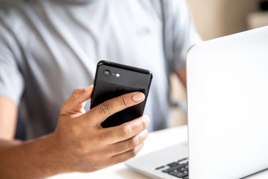 Ein Mann checkt sein Smartphone, während er am Laptop arbeitet: Über den WebAuthn-Standard kann das Smartphone auch als zweiter Faktor bei Log-ins am Rechner fungieren.