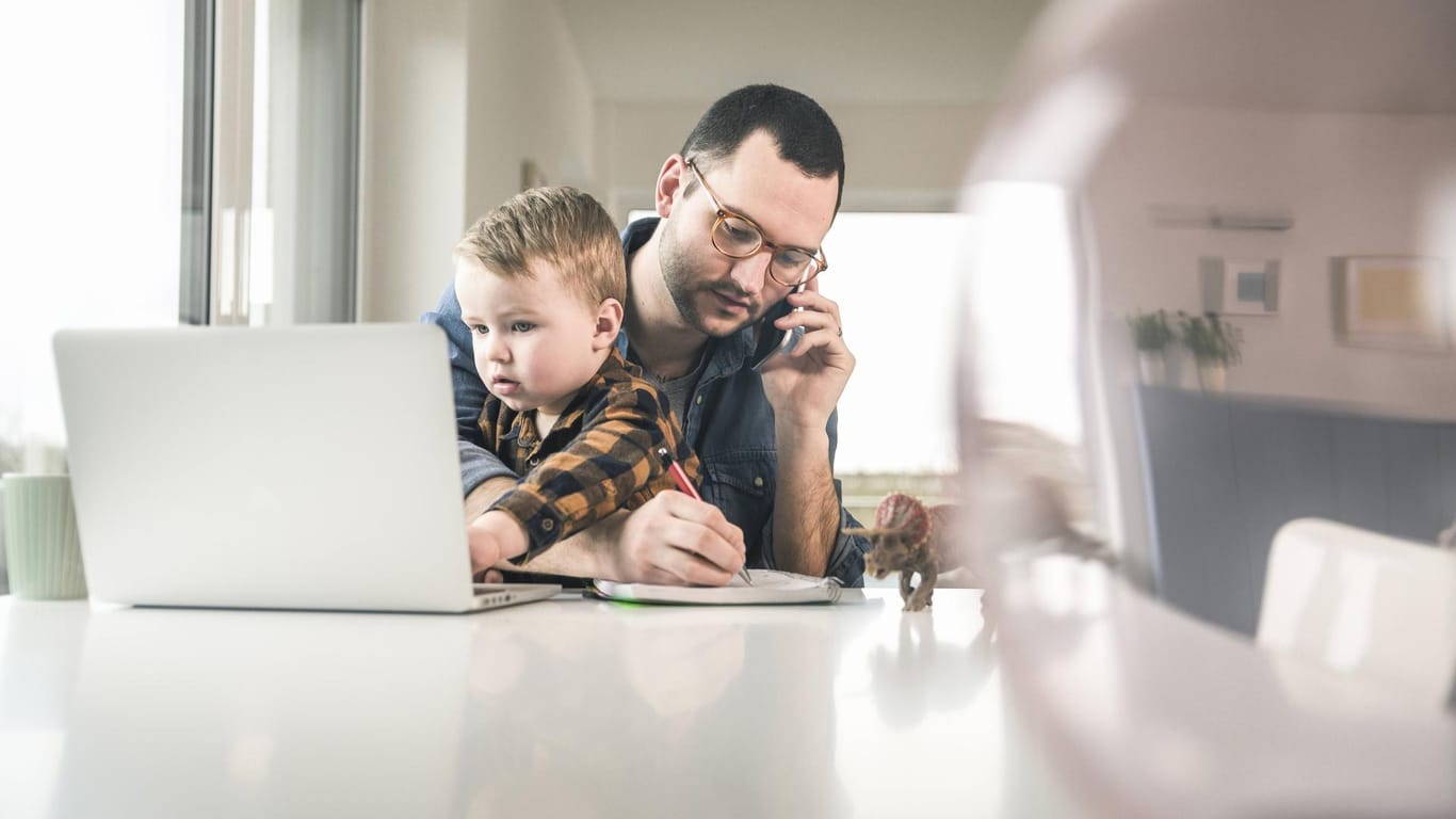 Ein Mann sitzt mit einem Notebook am Tisch, telefoniert und kümmert sich um seinen Sohn: Gleichzeitig mehrere Aufgaben zu erledigen, ist laut einer neuen Studie nicht sinnvoll.