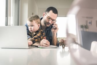 Ein Mann sitzt mit einem Notebook am Tisch, telefoniert und kümmert sich um seinen Sohn: Gleichzeitig mehrere Aufgaben zu erledigen, ist laut einer neuen Studie nicht sinnvoll.