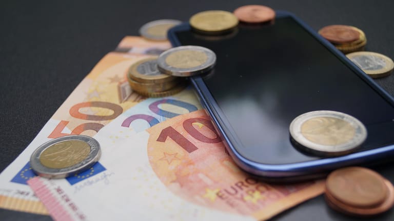 Ein Handy liegt inmitten von Euro-Münzen und -Scheinen: Ein neues Gesetz soll unter anderem die Laufzeit von Mobilfunkverträgen begrenzen.