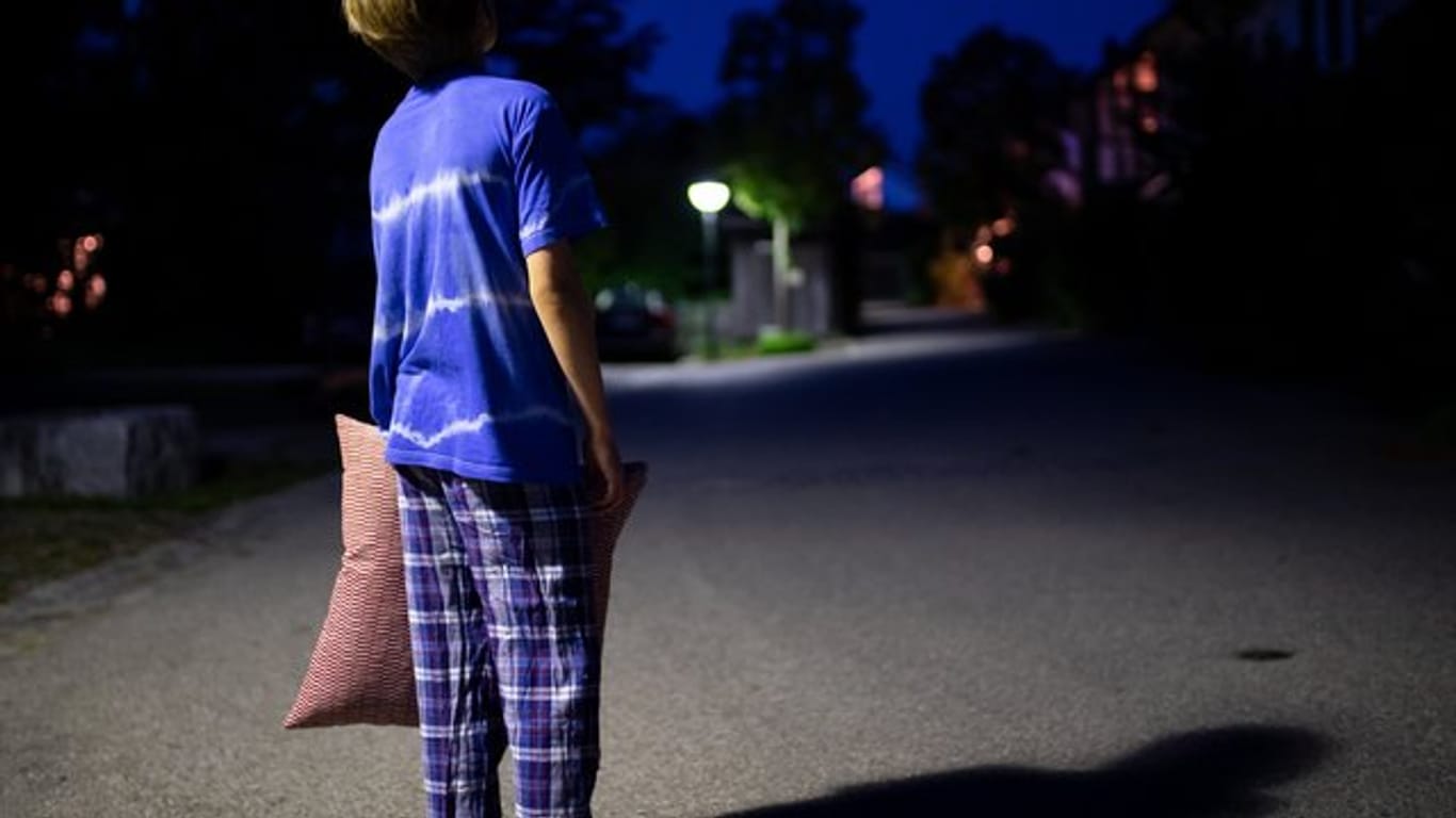 Im Schlafanzug steht ein Kind mit einem Kissen in der Hand nachts auf einer Straße.