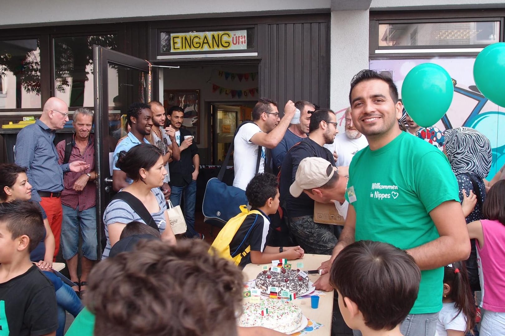 Menschen feiern mit Kuchen und Luftballons vor einem Gebäude: Fest zum zweijährigen Bestehen des benachbarten "Café international“.