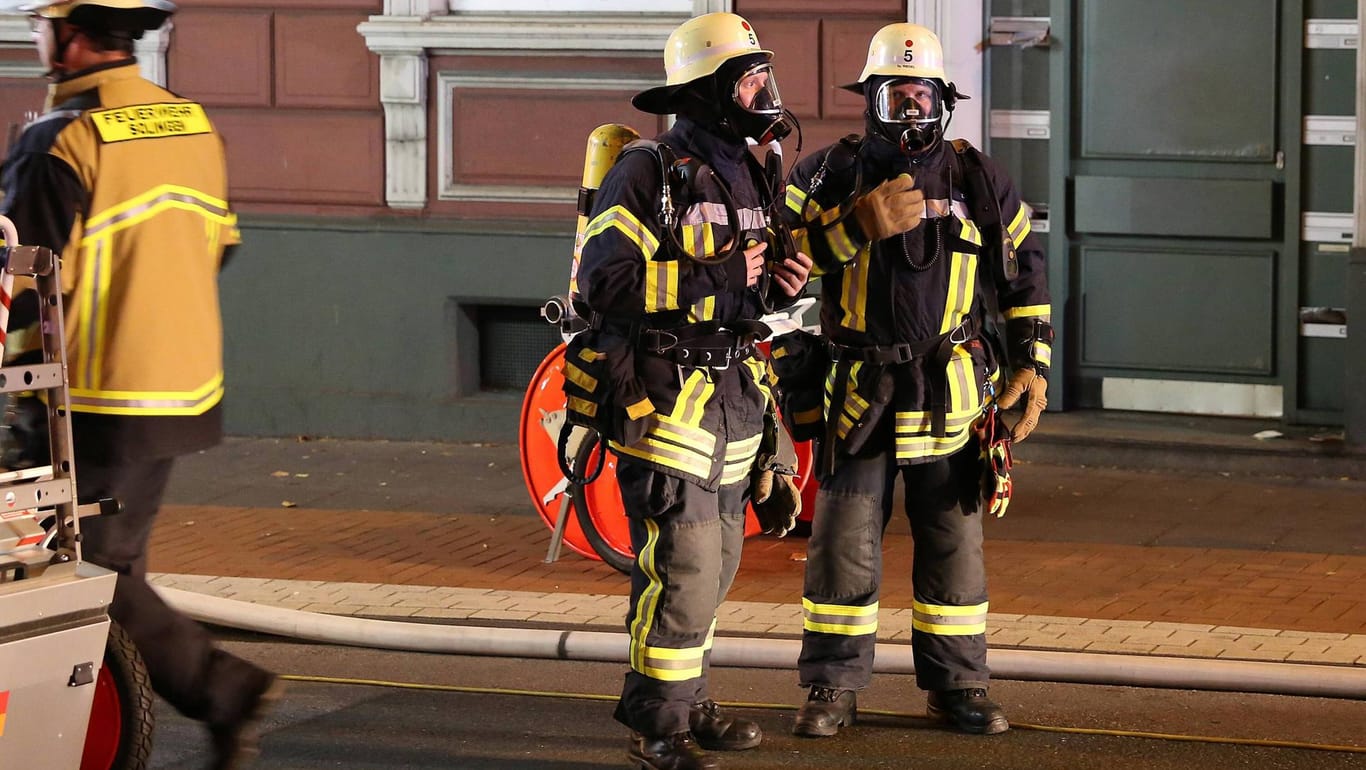 Zwei Feuerwehrmänner stehen in voller Montur vor einem Mehrfamilienhaus: In Wuppertal hat wieder ein Keller gebrannt.