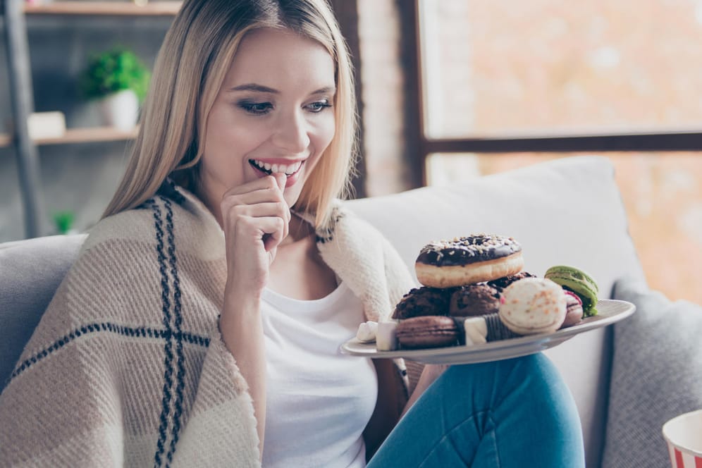 Eine Frau sitzt mit Kuchen auf dem Sofa: Am Cheat Day kann die hohe Zuckerzufuhr zu einer starken Erhöhung des Blutzuckerspiegels führen.
