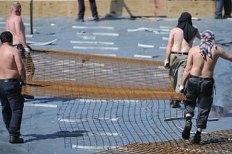 Arbeiter mit freiem Oberkörper tragen auf einer Baustelle ein Stahlgitter.