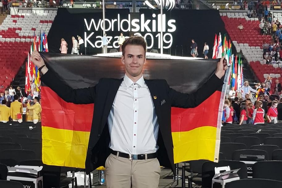 Ein Mann steht mit einer Deutschlandfahne in einer Halle: Der 19-Jährige ist ein wahres Talent im Programmieren.