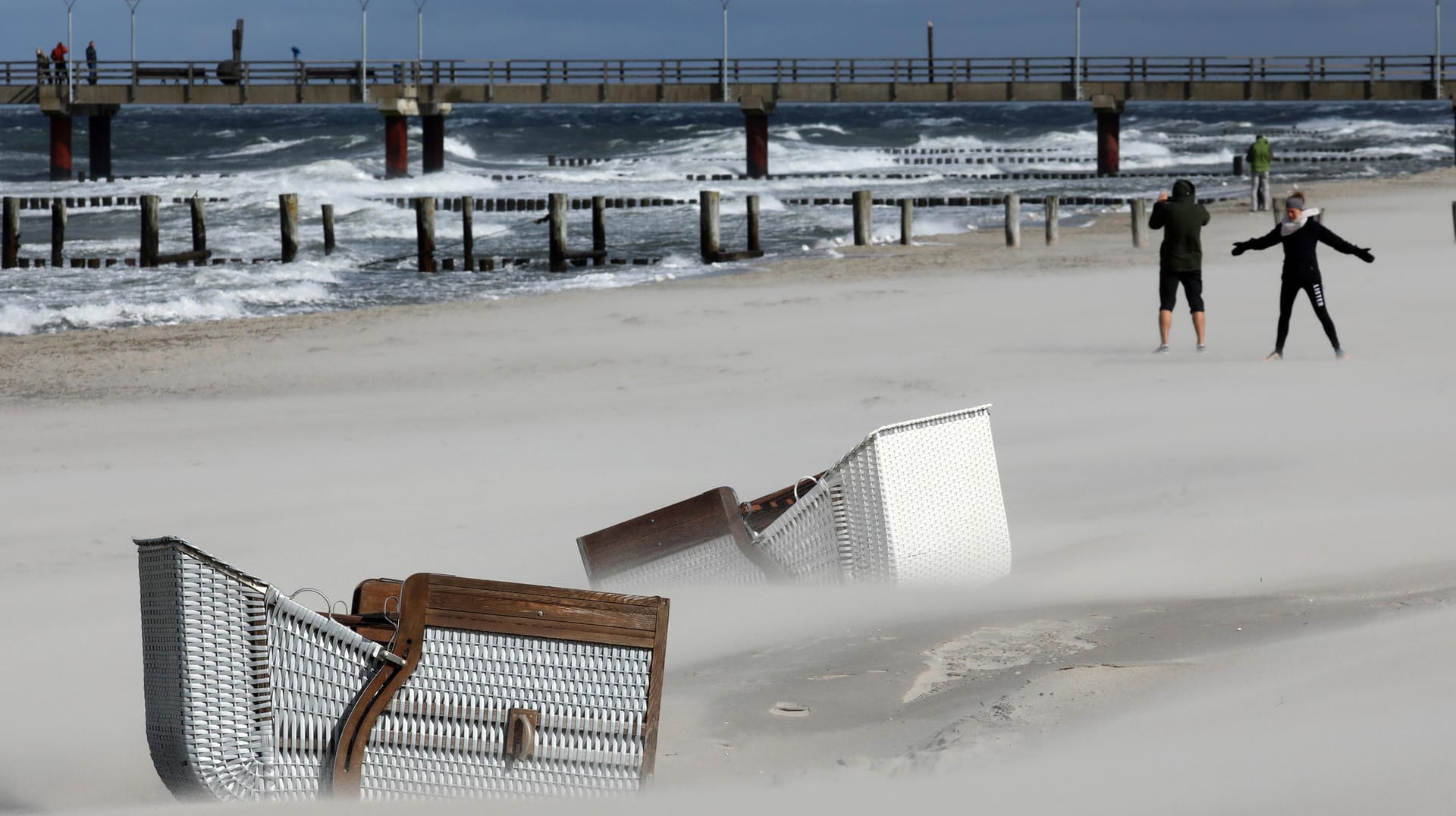 Zingst: Umgefallene Strandkörbe liegen im Sand, während sich Spaziergänger bei Sturm und Regen am Ostseestrand die Füße vertreten. Sonne und dunkle Wolken wechselten sich ständig ab.