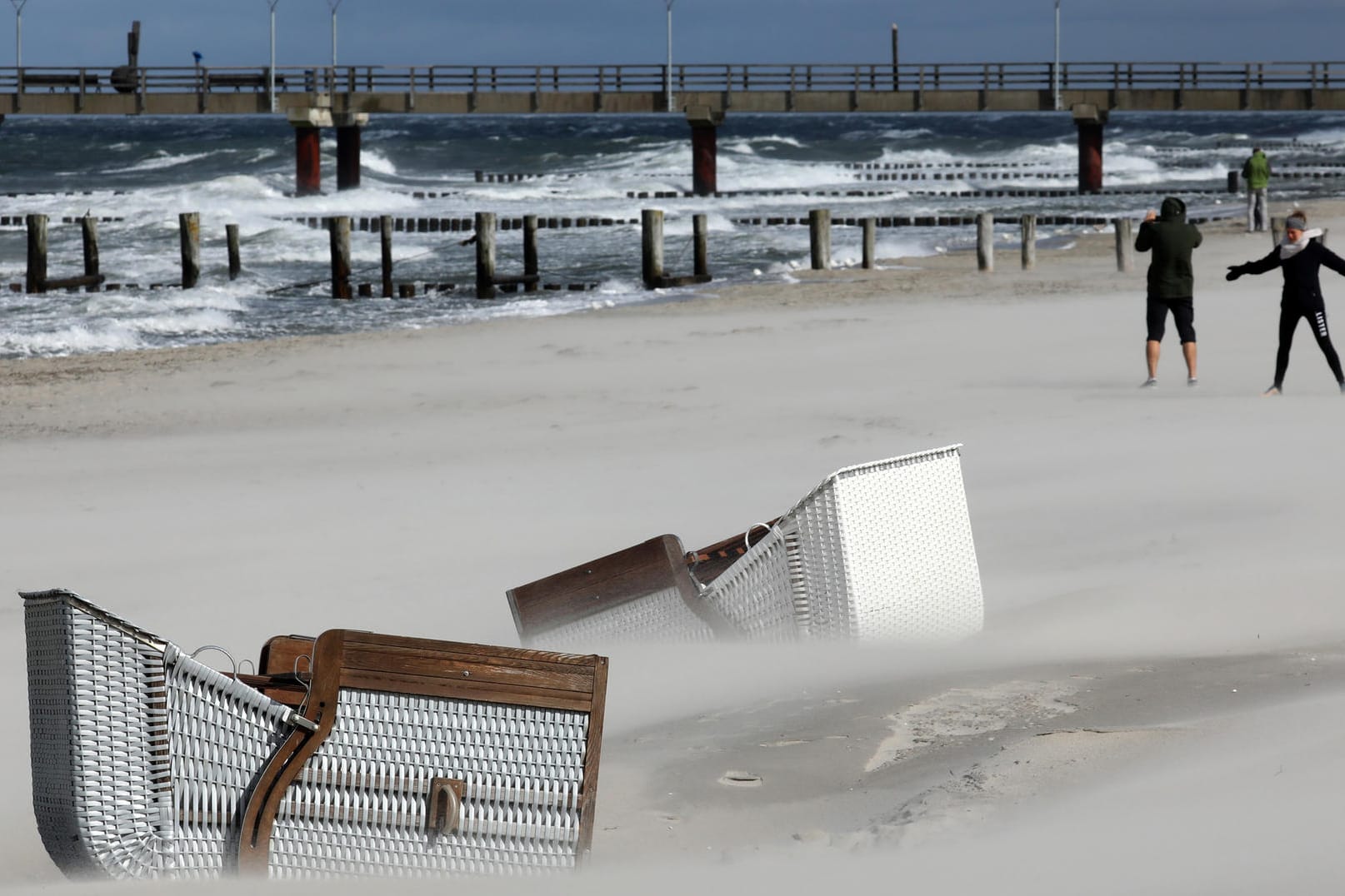 Zingst: Umgefallene Strandkörbe liegen im Sand, während sich Spaziergänger bei Sturm und Regen am Ostseestrand die Füße vertreten. Sonne und dunkle Wolken wechselten sich ständig ab.