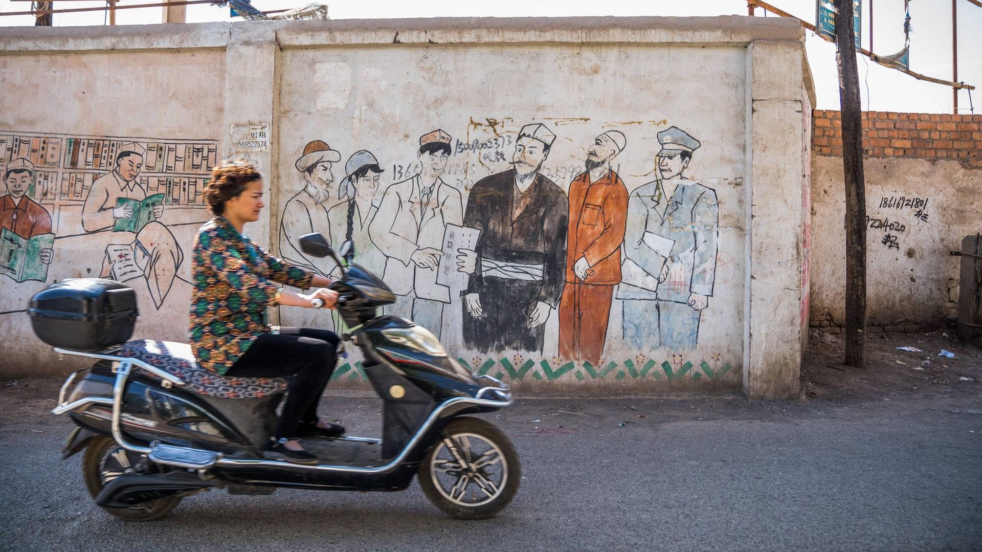 Wandbild in Turpan: Eine Frau fährt an einer chinesischen Propaganda-Darstellung in der Autonomen Region Xinjiang vorbei. (13. April 2019)