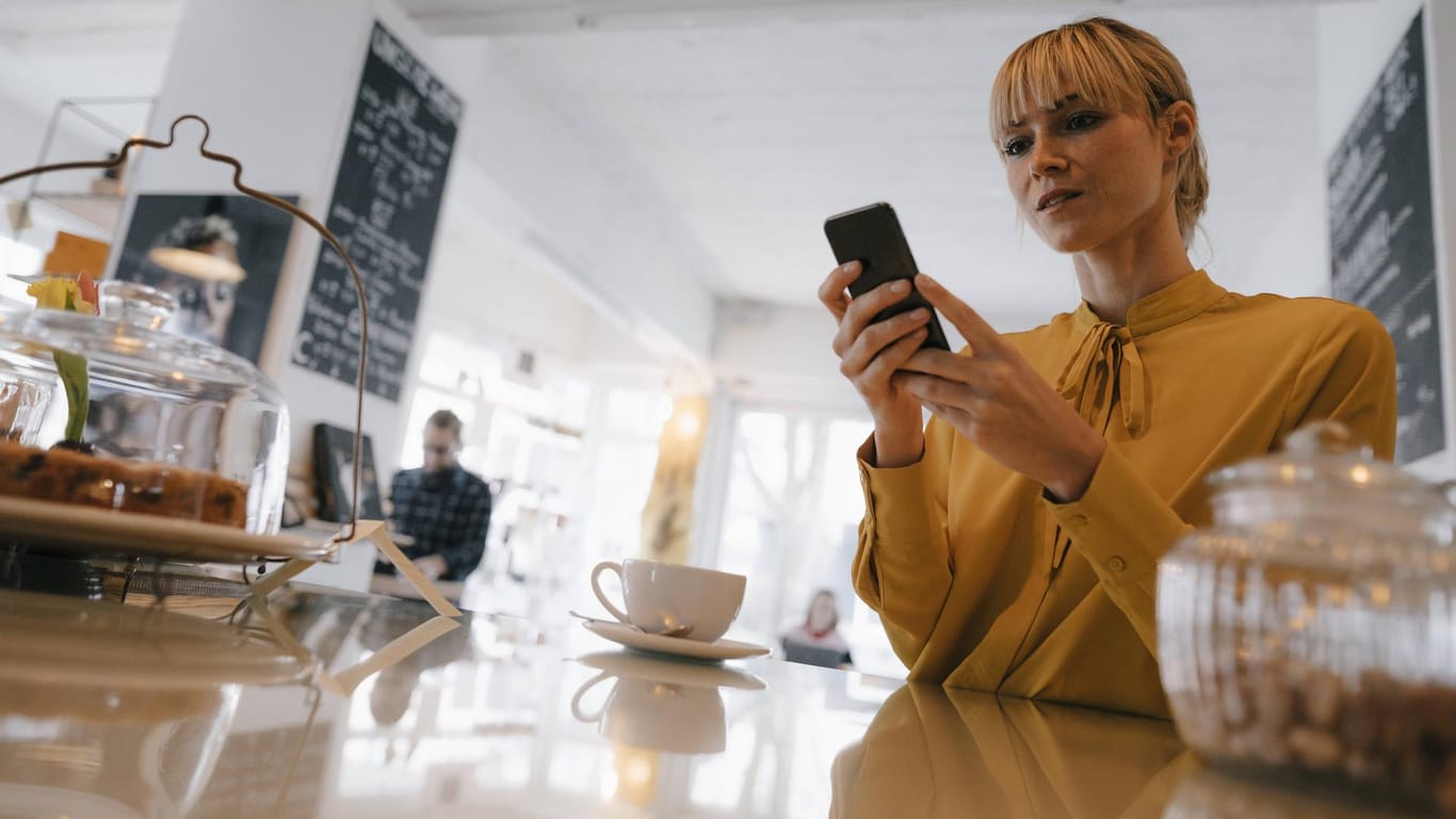 Eine junge Frau schaut im Café auf ihr Smartphone: Die Bundesnetzagentur hat Vodafone verboten, zu hohe Gebühren für die Telefonnummermitnahme zu kassieren.