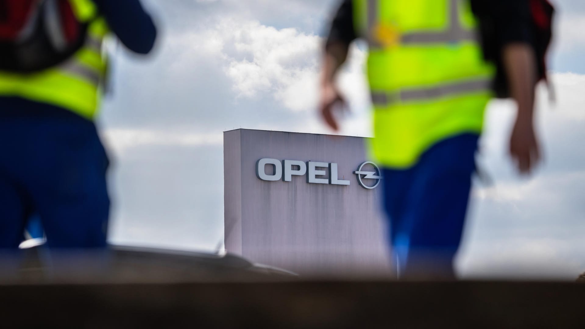 Zwei Männer in Sicherheitswesten gehen am Stammwerk von Opel vorbei: Der Autobauer Opel streicht in seinem Stammwerk Rüsselsheim für sechs Monate die Spätschicht.