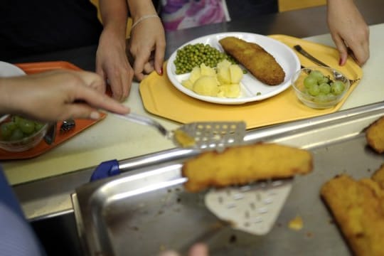 Eine Küchenhelferin legt an einer Gesamtschule ein Stück Fisch auf den Teller eines Schülers.