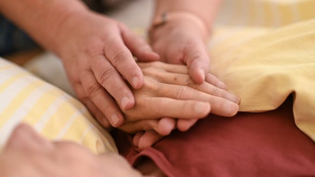 Eine Hospizmitarbeiterin hält die Hand eines todkranken Menschen, der im Hospiz im Bett liegt. Eine Hospizmitarbeiterin hält die Hand eines todkranken Menschen, der im Hospiz im Bett liegt.
