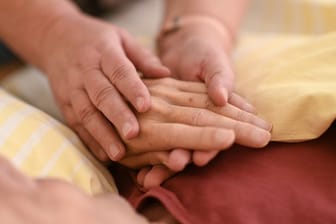 Eine Hospizmitarbeiterin hält die Hand eines todkranken Menschen, der im Hospiz im Bett liegt.