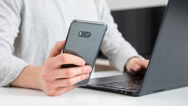 Ein Mann am Rechner mit Smartphone in der Hand: Ist das Internet ist zu langsam oder die Handyrechnung zu hoch, können Kunden ihre Rechte mit Hilfe von Musterbriefen einfordern.