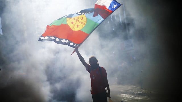 Ein vermummter Demonstrant weht die Flagge von Chile und der indigenen Gemeinschaft Mapuche mitten im Rauch während eines Protestes für Sozialreformen. Ein vermummter Demonstrant weht die Flagge von Chile und der indigenen Gemeinschaft Mapuche mitten im Rauch während eines Protestes für Sozialreformen.