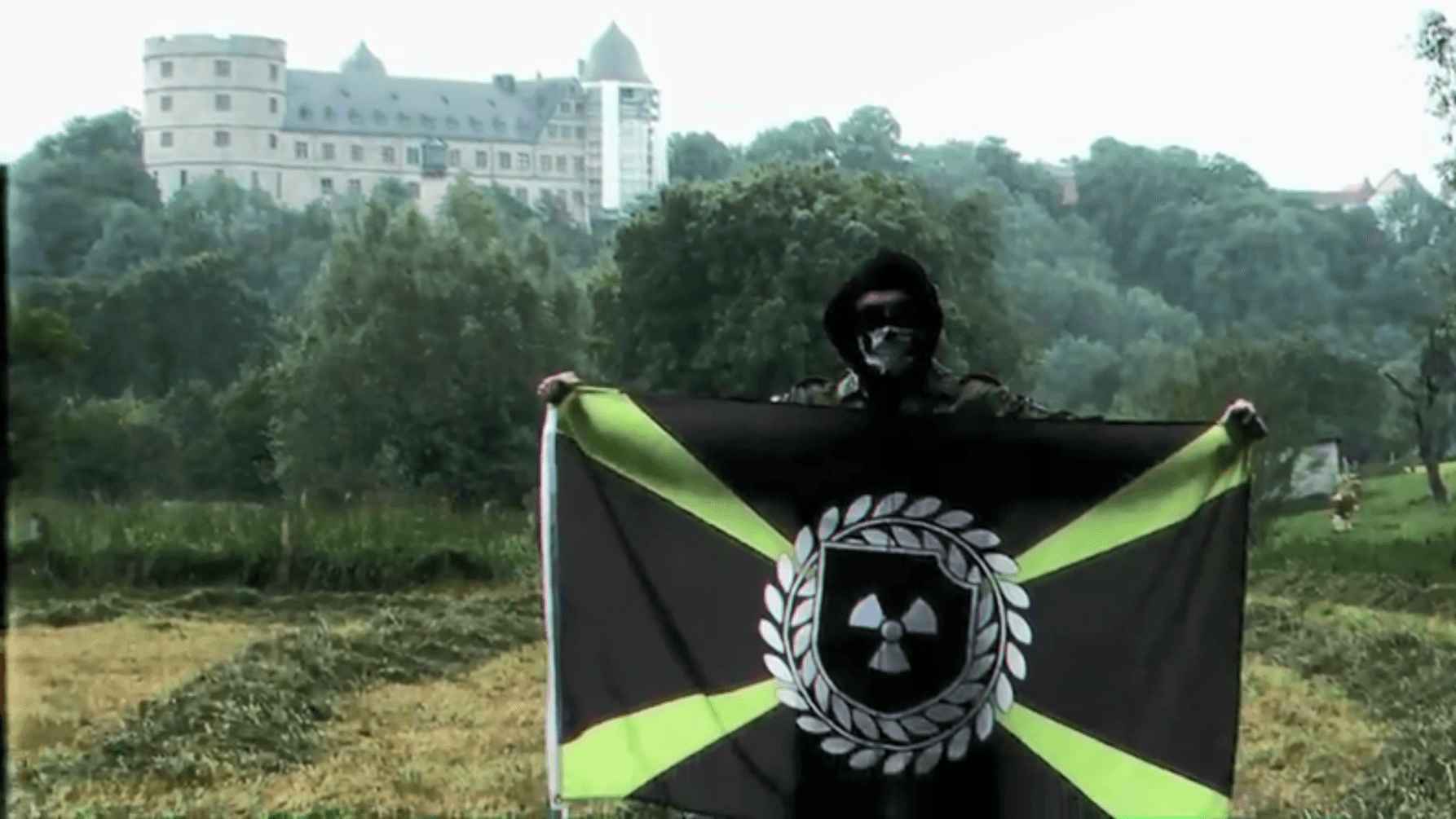 "Atomwaffen Division": Ein Neonazi mit der Flagge der Organisation steht im einzigen Video der deutschen Organisation vor der Wewelsburg. Ein Datenleck gibt Hinweise zu einem möglichen Beteiligten aus Thüringen.