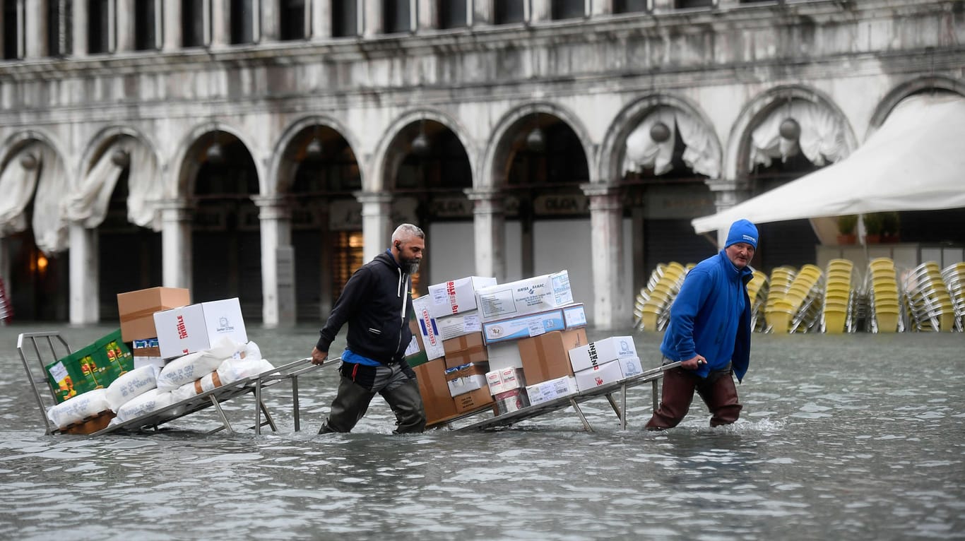 Männer waten mit Kartons und Lebensmitteln, die sie auf einer Trage transportieren, durch das Hochwasser auf einem überfluteten Platz.