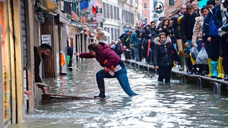 Eine Frau versucht, eine überflutete Straße zu überqueren, während Menschen bei Hochwasser auf einer Gerüstbrücke laufen.