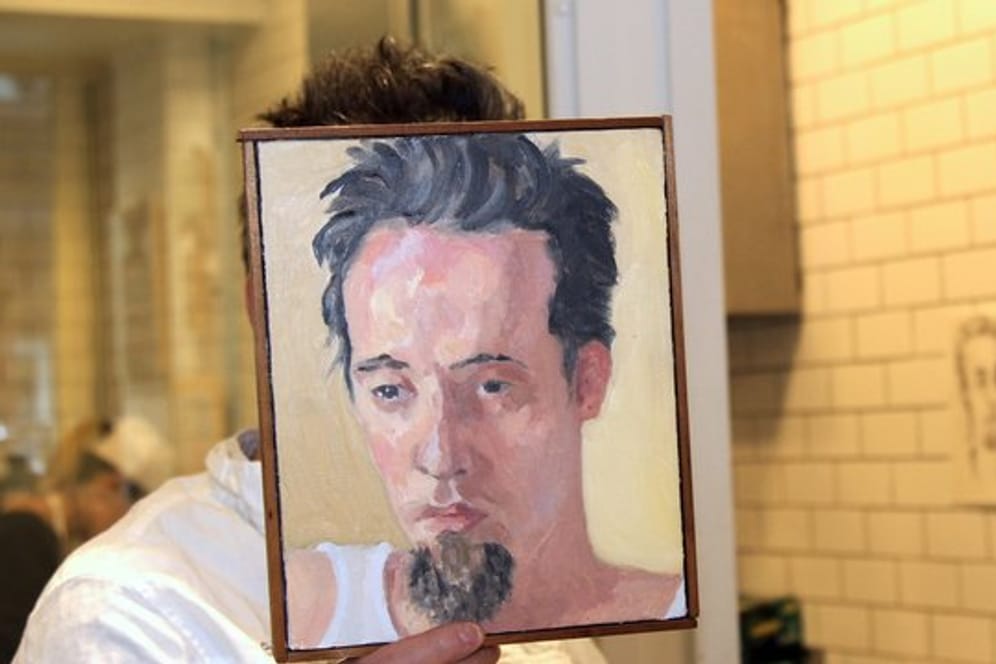 Sebastian Brecht hält sich ein Portrait vor sein Gesicht, das die Künstlerin Alix Bailey von ihm gemalt hat.