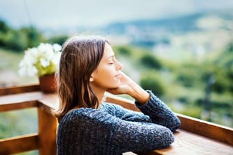 Eine Frau steht mit geschlossenen Augen auf einem Balkon: Kleine Pausen im Alltag sind auch eine Form der Selbstfürsorge.