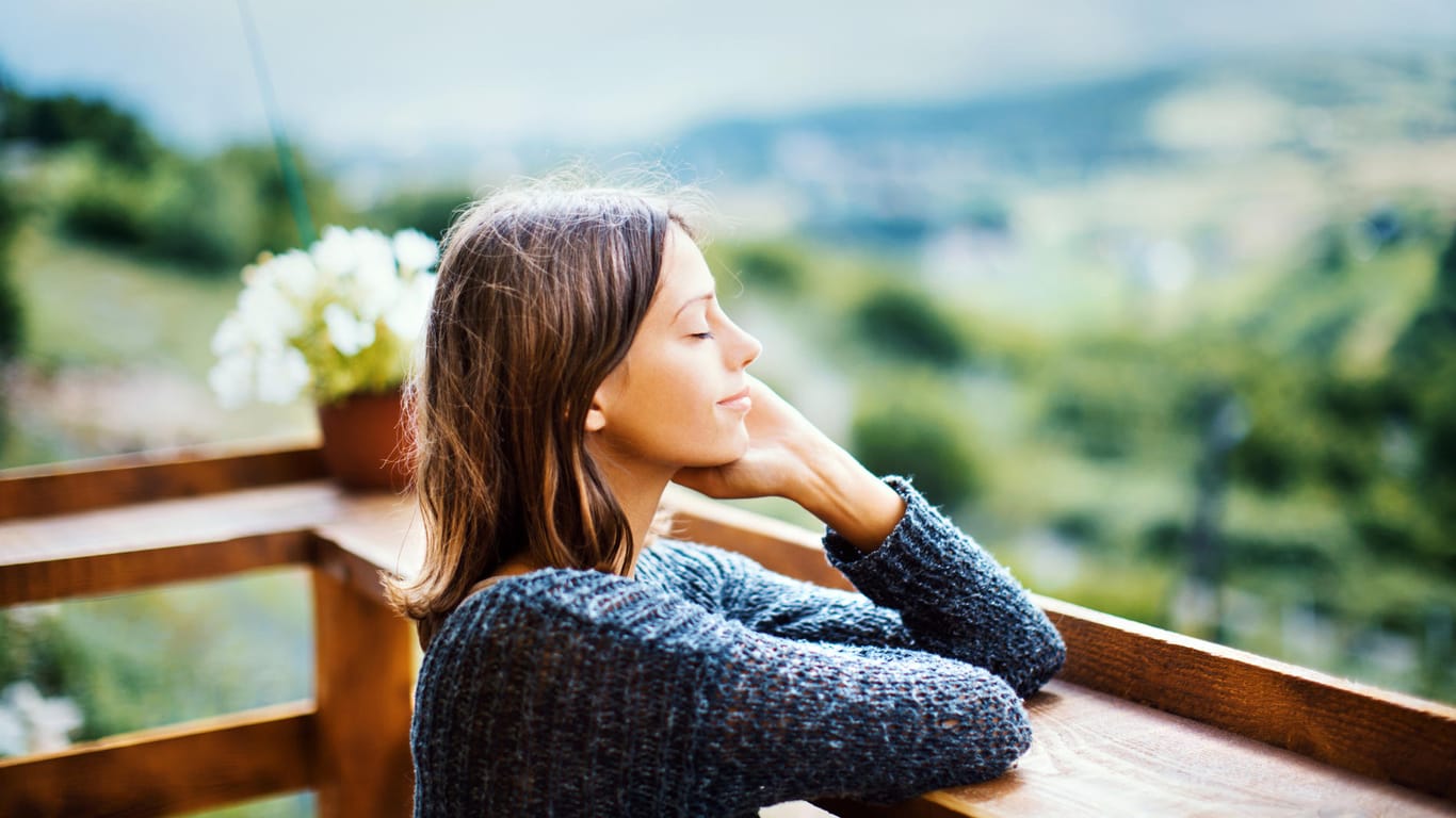 Eine Frau steht mit geschlossenen Augen auf einem Balkon: Kleine Pausen im Alltag sind auch eine Form der Selbstfürsorge.