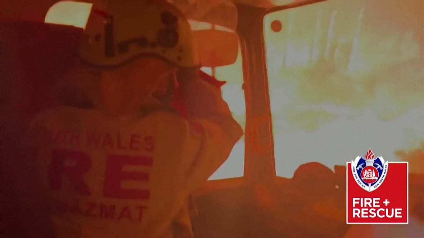 Die Flammen umgeben den Feuerwehrtruck: Mit einer Decke versuchen die Feuerwehrmänner sich gegen die Hitze zu schützen.