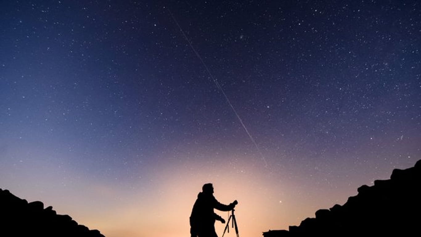 Ein Fotograf steht vor einem Nachthimmel, an dem die Bewegung von Starlink-Satelliten als leuchtende Spur (Bildmitte) sichtbar wird.