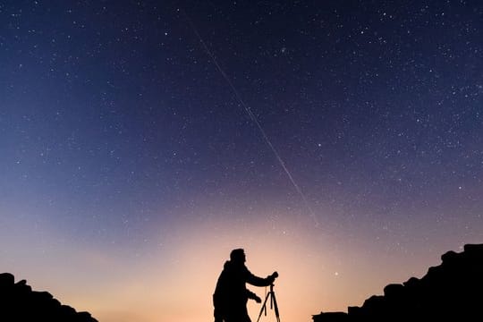 Ein Fotograf steht vor einem Nachthimmel, an dem die Bewegung von Starlink-Satelliten als leuchtende Spur (Bildmitte) sichtbar wird.