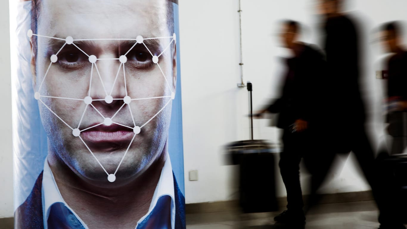 Menschen laufen an einem Poster mit schematischer Darstellung von Gesichtserkennung vorbei: Die Technik zur automatischen Gesichtserkennung ist umstritten.