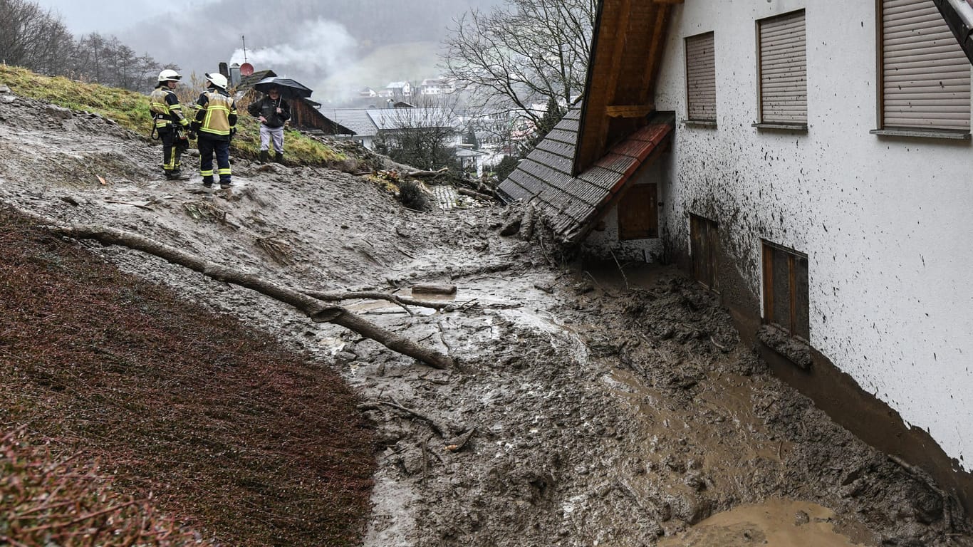Simonswald in Baden-Württemberg: Feuerwehrmänner stehen vor an einem Haus, das von einer Schlammlawine getroffen wurde.