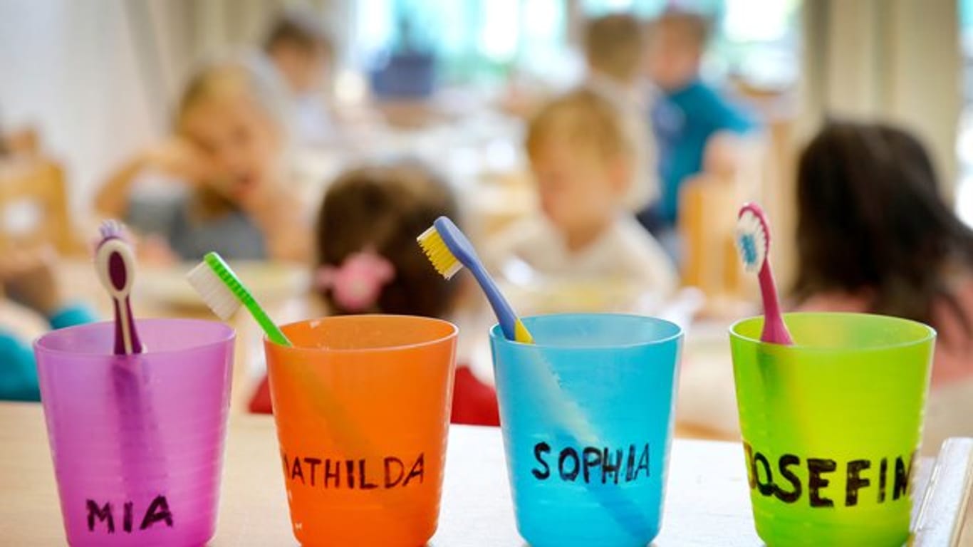 Zahnbürsten und Zahnputzbecher mit den Vornamen der Kinder stehen in einer Kindertagesstätte auf einem Tisch.