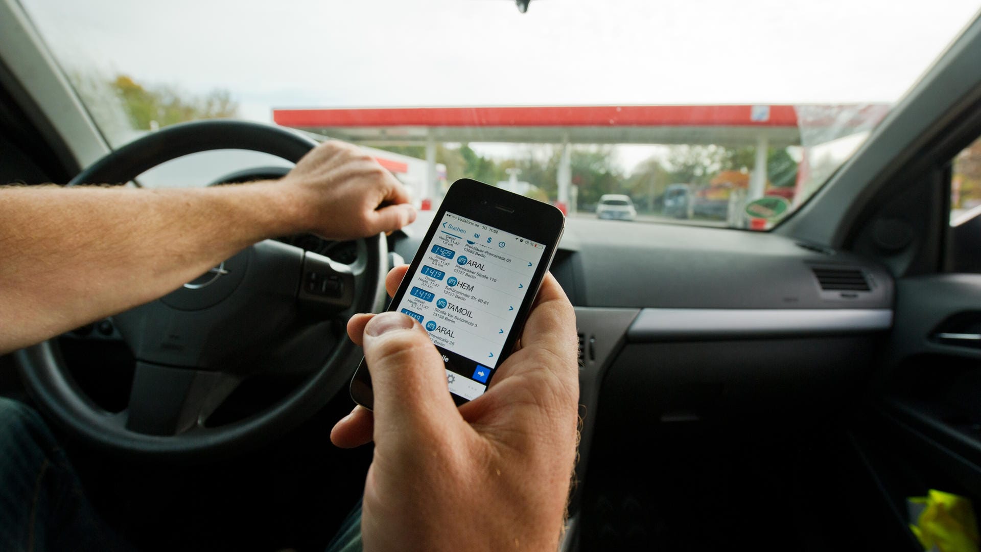 Ein Mann sitzt in einem Auto und überprüft mit einem Smartphone die aktuellen Benzinpreise.