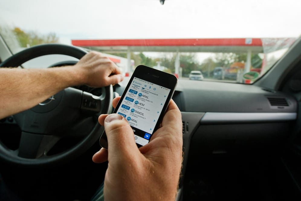 Ein Mann sitzt in einem Auto und überprüft mit einem Smartphone die aktuellen Benzinpreise.