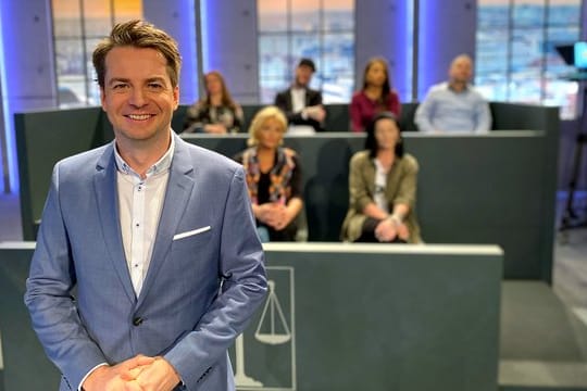 Alexander Stevens, Moderator und juristischer Experte, steht vor den sieben Juroren.