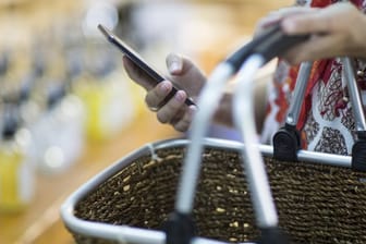 Eine Frau hält einen Einkaufskorb und ihr Smartphone (Symbolbild): In Mainz bieten Freiwillige an, für Risikogruppen einkaufen zu gehen.