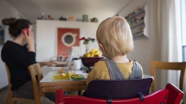 Eine Frau sitzt im Homeoffice an ihrem Laptop und telefoniert, während ihr Kind neben ihr in einem Kinderstuhl am Tisch sitzt.