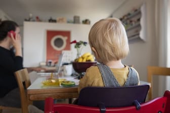 Eine Frau sitzt im Homeoffice an ihrem Laptop und telefoniert, während ihr Kind neben ihr in einem Kinderstuhl am Tisch sitzt.