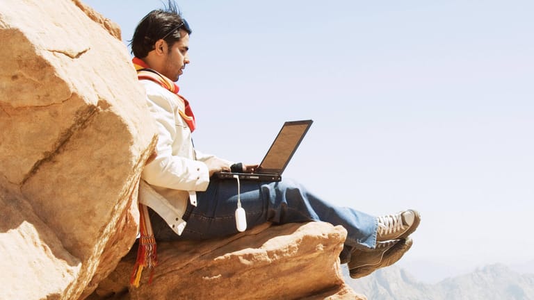 Ein Mann sitzt mit einem Laptop auf einem Felsen: So einfach können Sie davon profitieren, wenn ein Unternehmen einen Teil seines Gewinns ausschüttet.