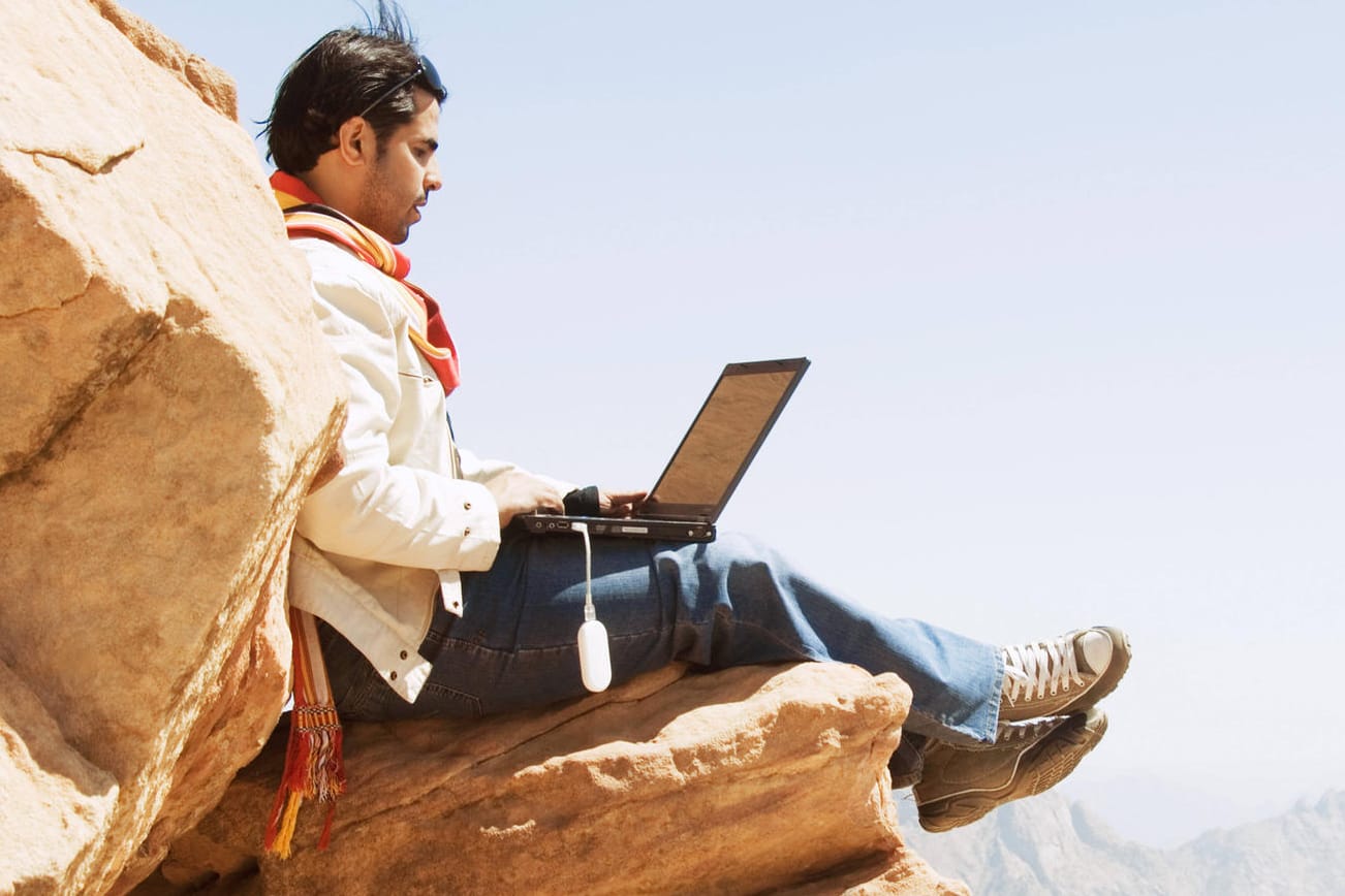 Ein Mann sitzt mit einem Laptop auf einem Felsen: So einfach können Sie davon profitieren, wenn ein Unternehmen einen Teil seines Gewinns ausschüttet.