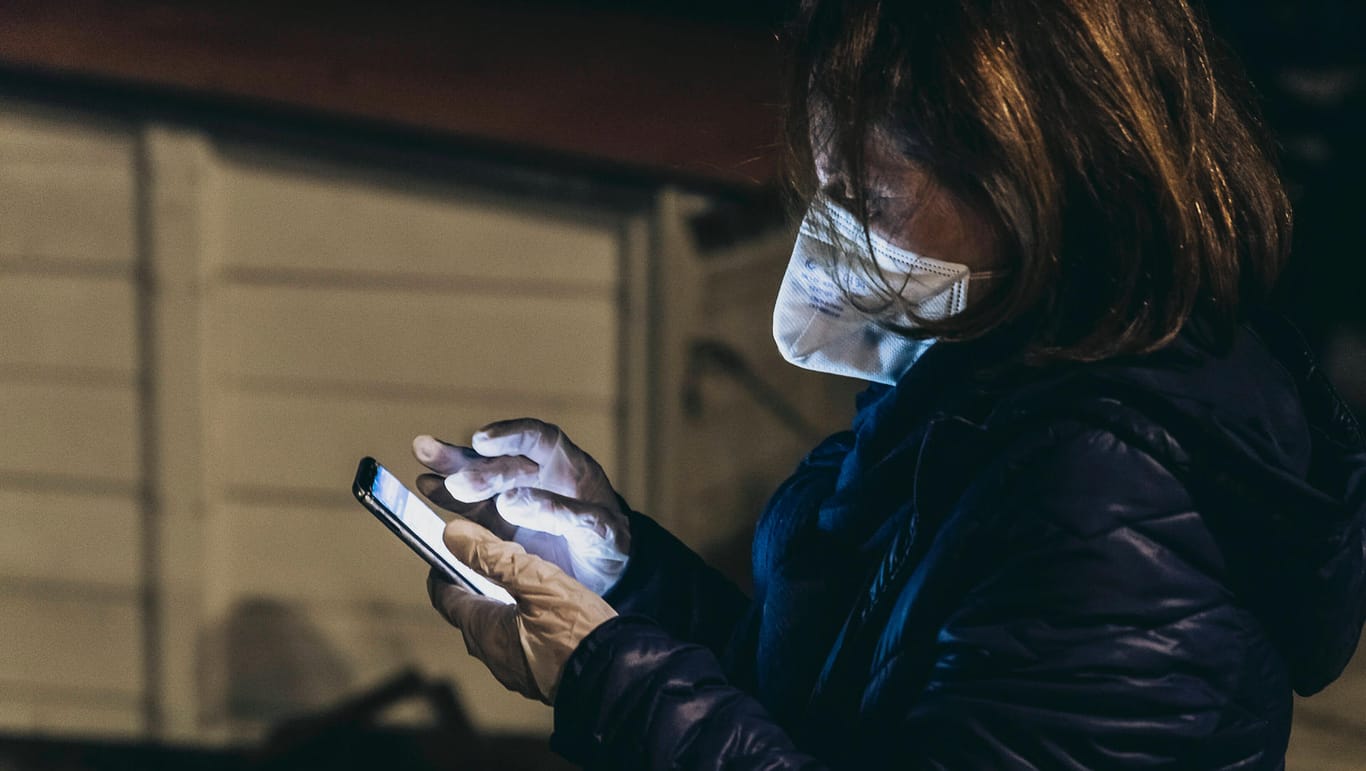 Eine Frau mit Atemschutzmaske und Handschuhen tippt in ein Smartphone: Bürger in Hagen können die Corona-Hotline nun noch länger erreichen (Symbolbild).