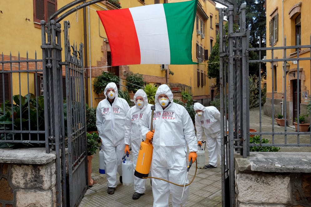 Arbeiter in Schutzkleidung desinfizieren Gehwege in Rom: Die Corona-Krise hat viele Länder hart getroffen, insbesondere Italien. Das stellt auch die Europäische Gemeinschaft auf die Probe – der Blick richtet sich vor allem auf Deutschland.