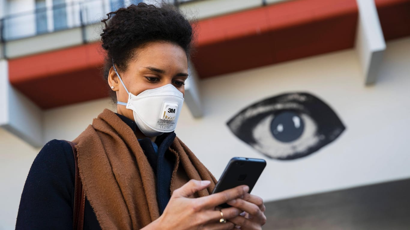 Eine Frau mit Smartphone und Gesichtsschutz (Symbolbild): Derzeit wird an einer App zur Analyse der Corona-Infektionswege gearbeitet.