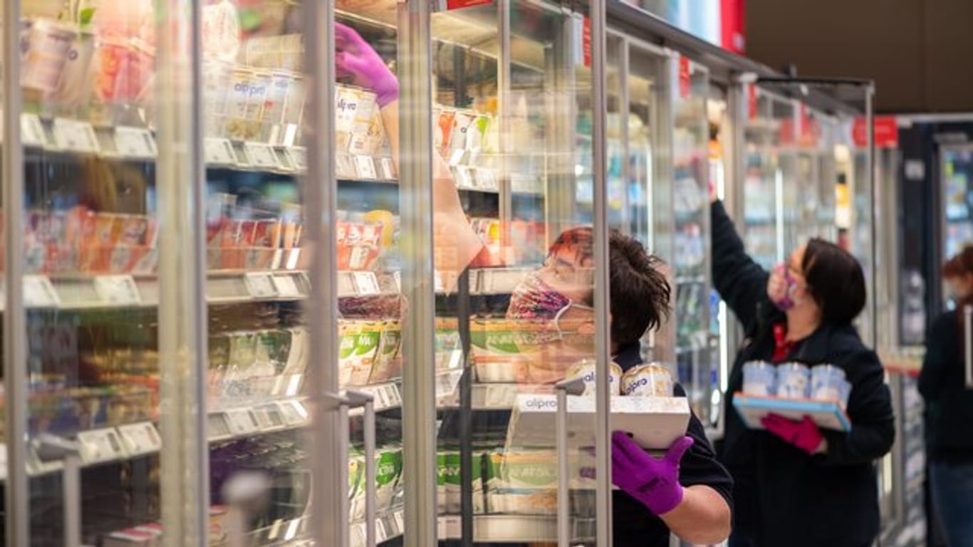 Mitarbeiterinnen in einem Supermarkt tragen Schutzmasken, während sie die Kühlregale mit Waren bestücken.